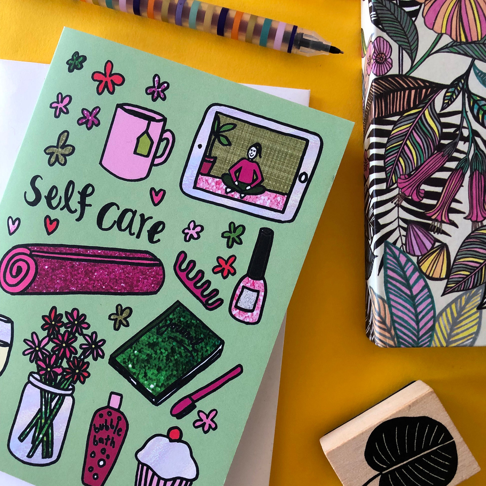 Image show a green greetings card that says 'self care' and features a fun pattern of self care items