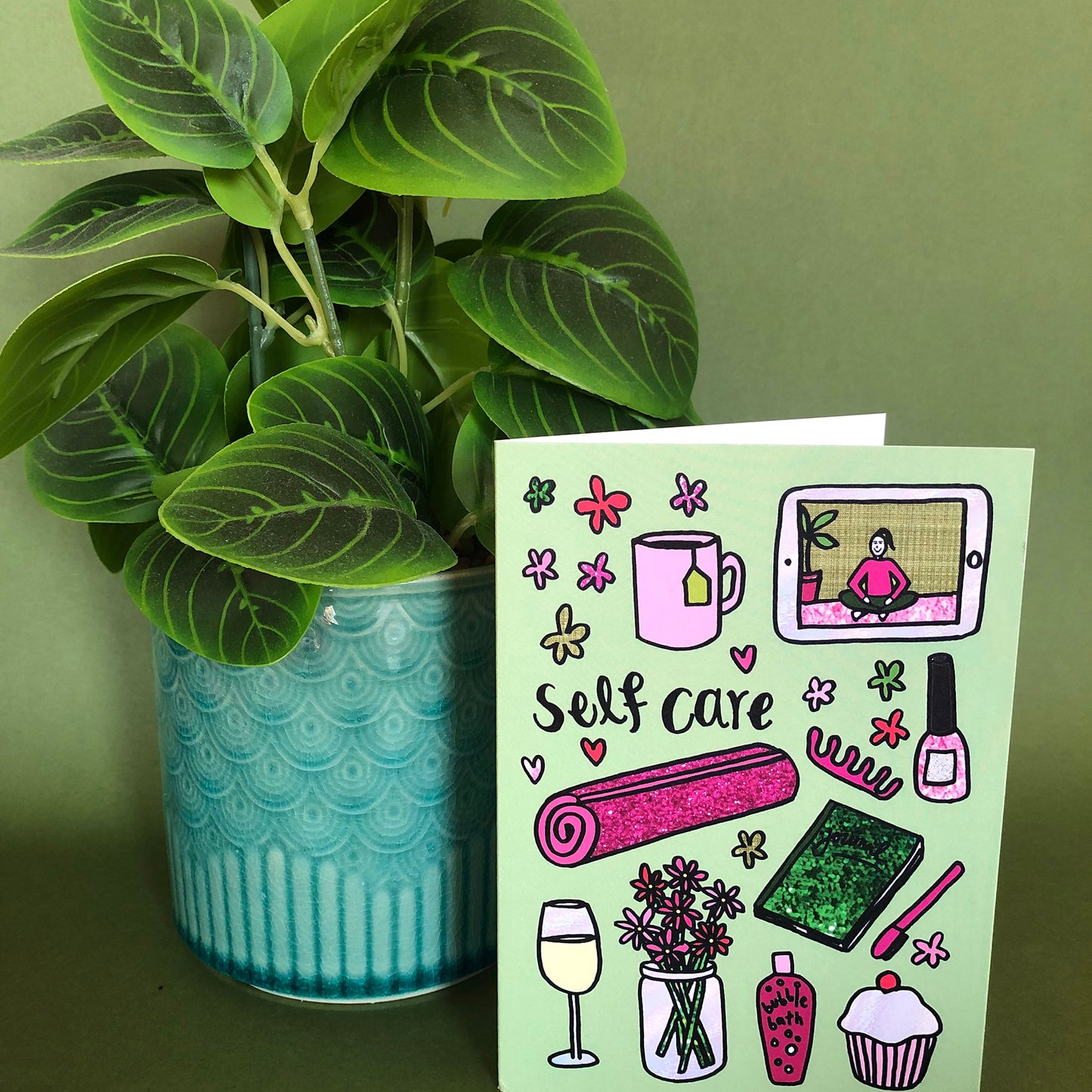 Image show a green greetings card that says 'self care' and features a fun pattern of self care items