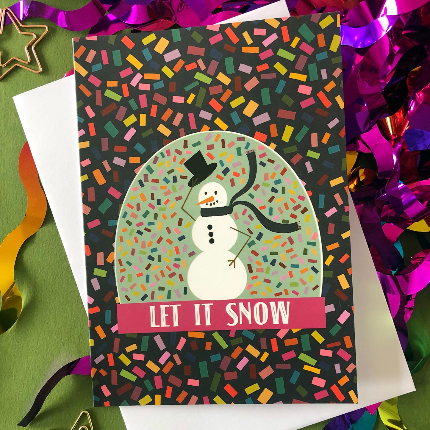 Image shows a bright and colourful Christmas card featuring an illustration of a snow globe on a confetti patterned background.