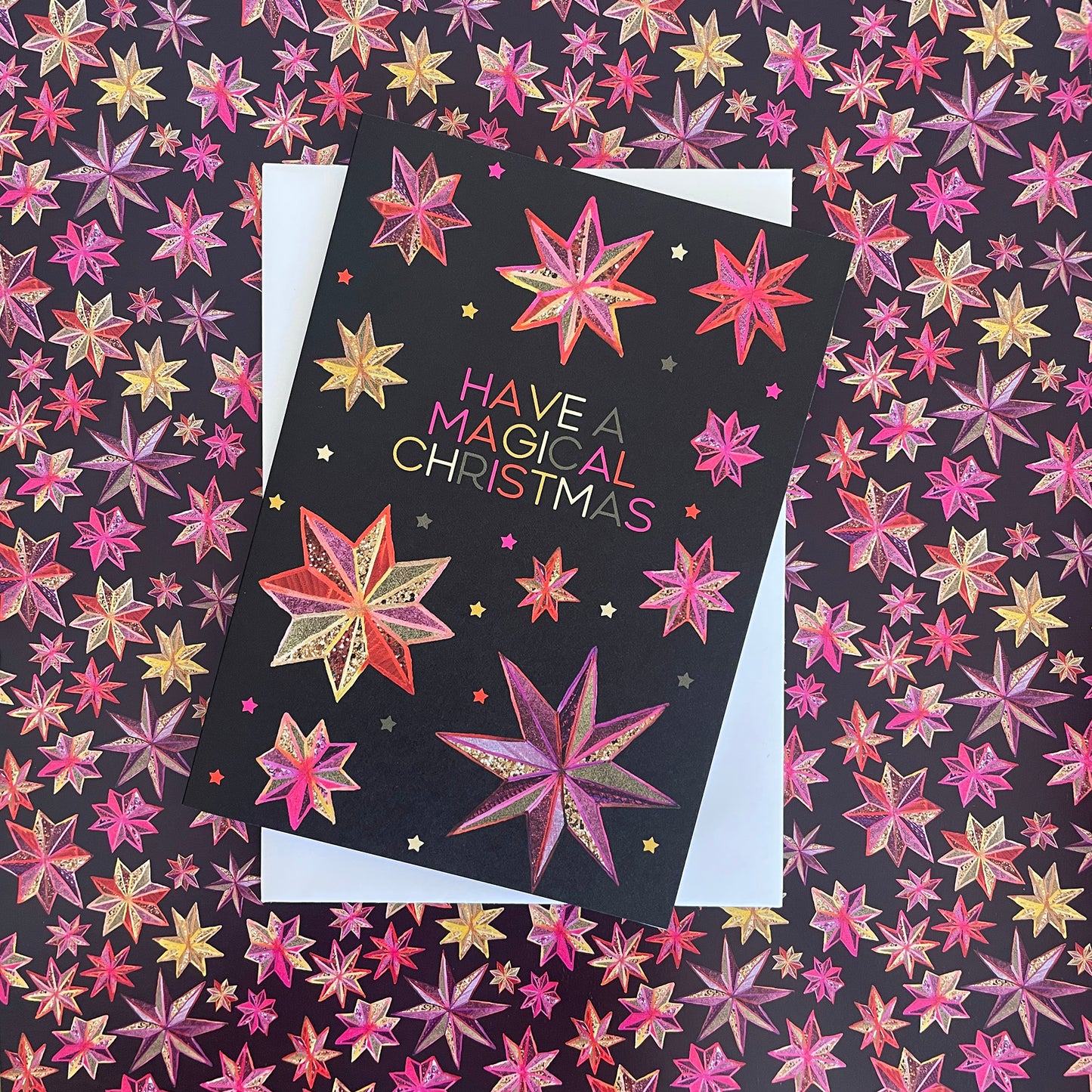 Image shows a black Christmas card with a hand drawn star pattern in pinks, oranges and golds with the wording 'Have a magical Christmas'