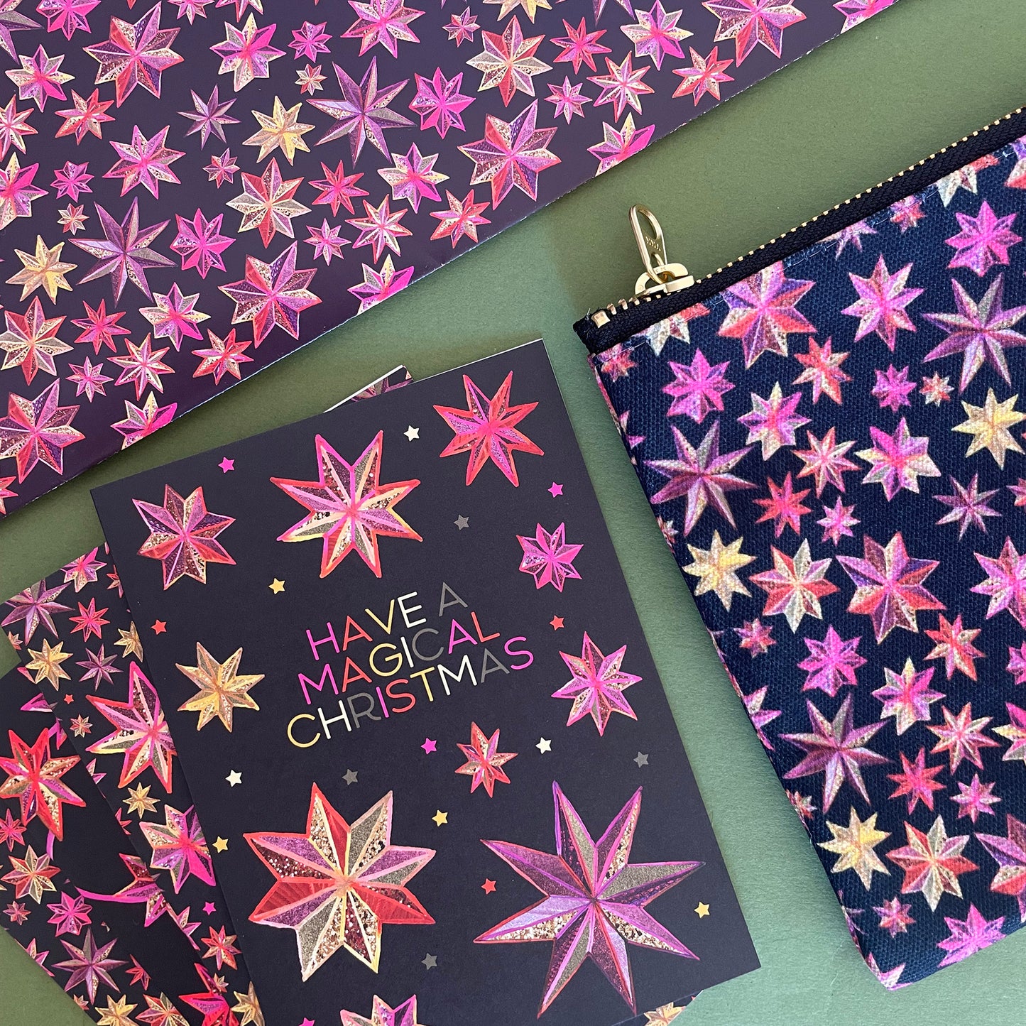 Image shows a black Christmas card with a hand drawn star pattern in pinks, oranges and golds with the wording 'Have a magical Christmas' along with matching gift wrap and zip pouch