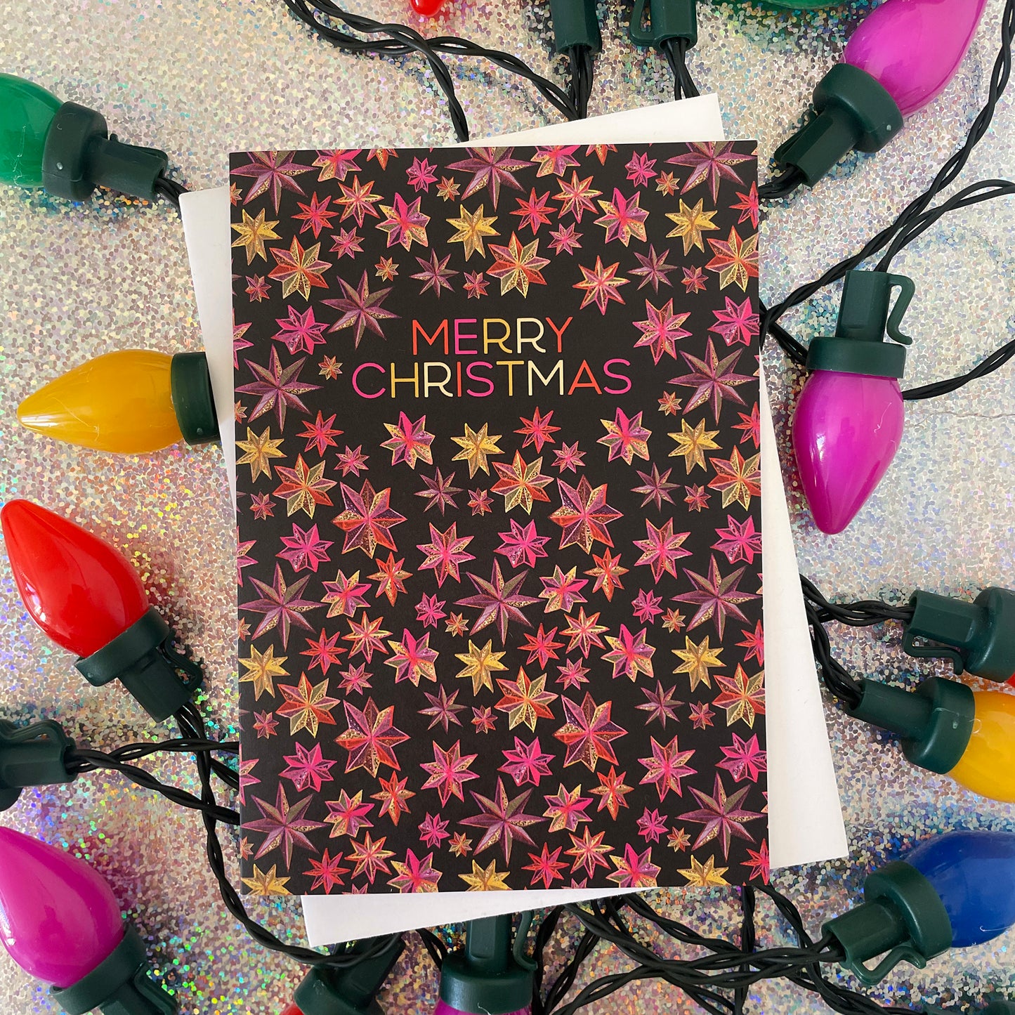 Image shows a black Christmas card with a hand drawn star pattern in pinks, oranges and golds with the wording 'Merry Christmas'