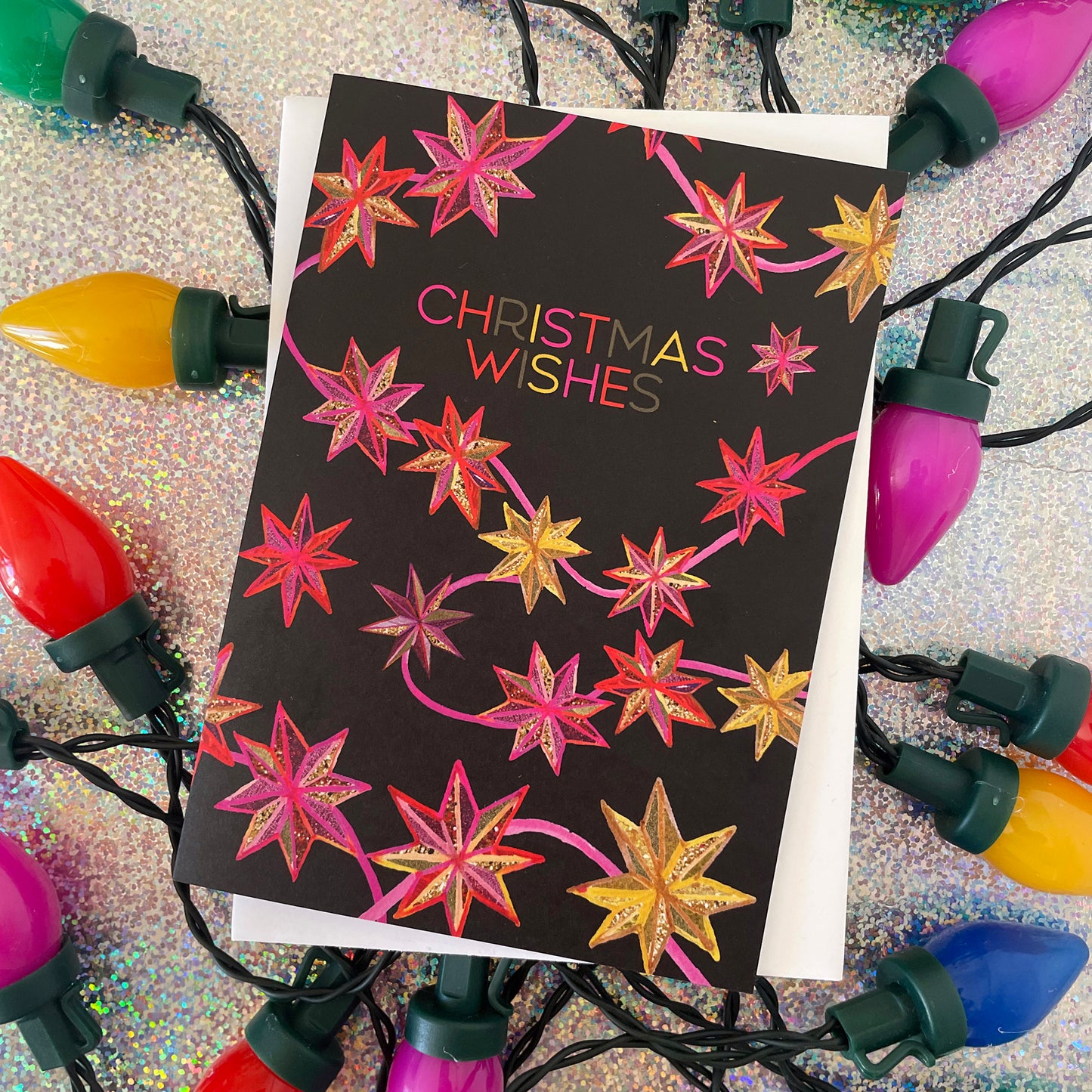 Image shows a black Christmas card with a hand drawn star pattern in pinks, oranges and golds with the wording 'Christmas Wishes'