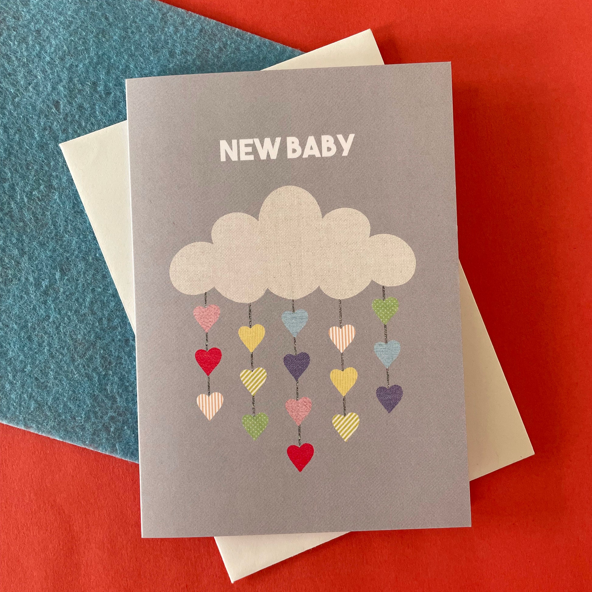 Image shows a pale grey greetings card with an illustration of a cloud with colourful hearts hanging from it and the words 'New Baby'