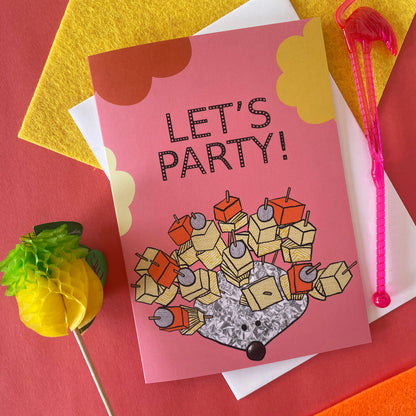 Image shows a pink greetings card with an illustration of a silver foil 'party hedgehog' with cheese, pineapple and pickled onions on cocktail sticks and the words 'let's party'