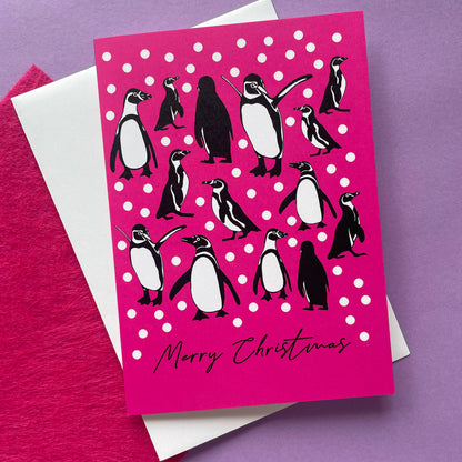 A Christmas greeting card featuring penguin illustrations on a bright pink background with white snow balls and the text 'Merry Christmas' at the bottom, styled with a white envelope and textured pink paper on a purple surface.