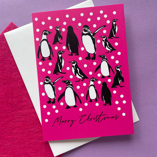 A Christmas greeting card featuring penguin illustrations on a bright pink background with white snow balls and the text 'Merry Christmas' at the bottom, styled with a white envelope and textured pink paper on a purple surface.