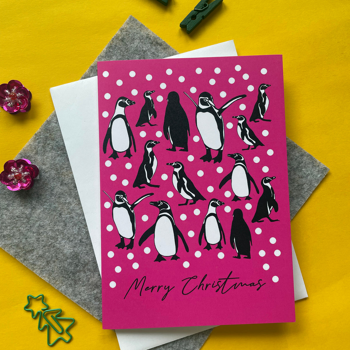 A Christmas greeting card featuring a pink background with white snowballs and illustrated penguins, placed on top of a grey felt envelope and white paper. The card reads 'Merry Christmas' and is photographed on a bright yellow surface with small decorative props.