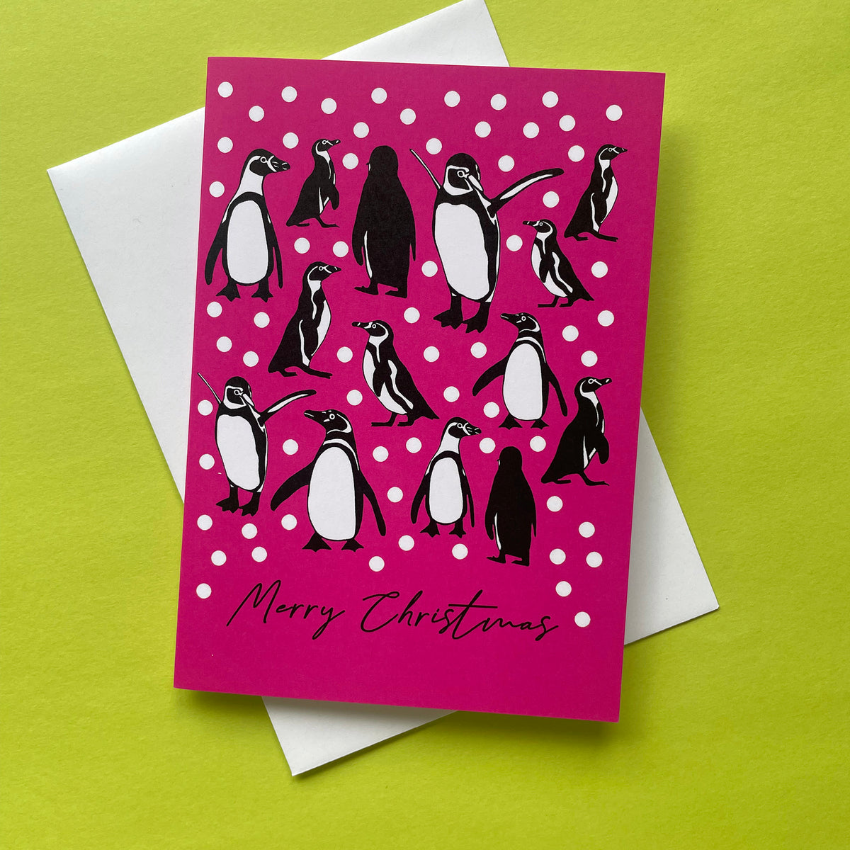 A Christmas greeting card with a bright pink background, white snowballs, and penguin illustrations, placed on top of a white envelope against a lime green background.