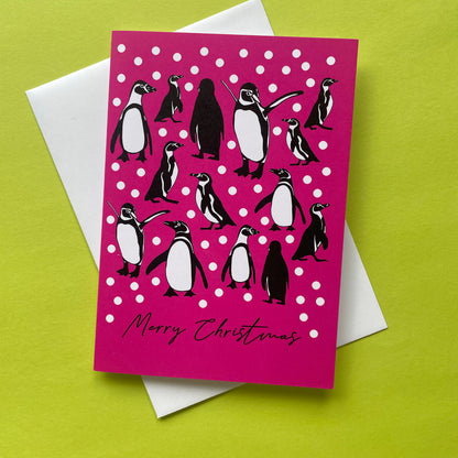 A Christmas greeting card with a bright pink background, white snowballs, and penguin illustrations, placed on top of a white envelope against a lime green background.