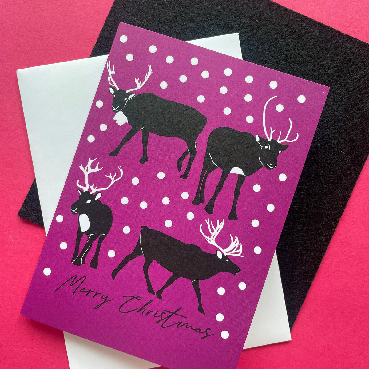 A Christmas greeting card with a magenta-purple background, white snowballs, and black reindeer silhouettes, layered over a white envelope and a black textured card on a bright pink background.