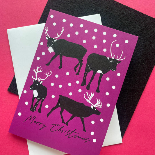 A Christmas greeting card with a magenta-purple background, white snowballs, and black reindeer silhouettes, layered over a white envelope and a black textured card on a bright pink background.