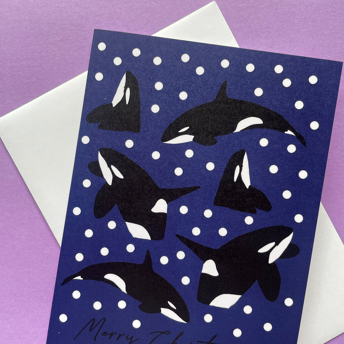 A Christmas greeting card featuring black and white orca illustrations on a dark blue background with white snowballs, shown with a white envelope on a purple surface.