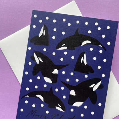 A Christmas greeting card featuring black and white orca illustrations on a dark blue background with white snowballs, shown with a white envelope on a purple surface.