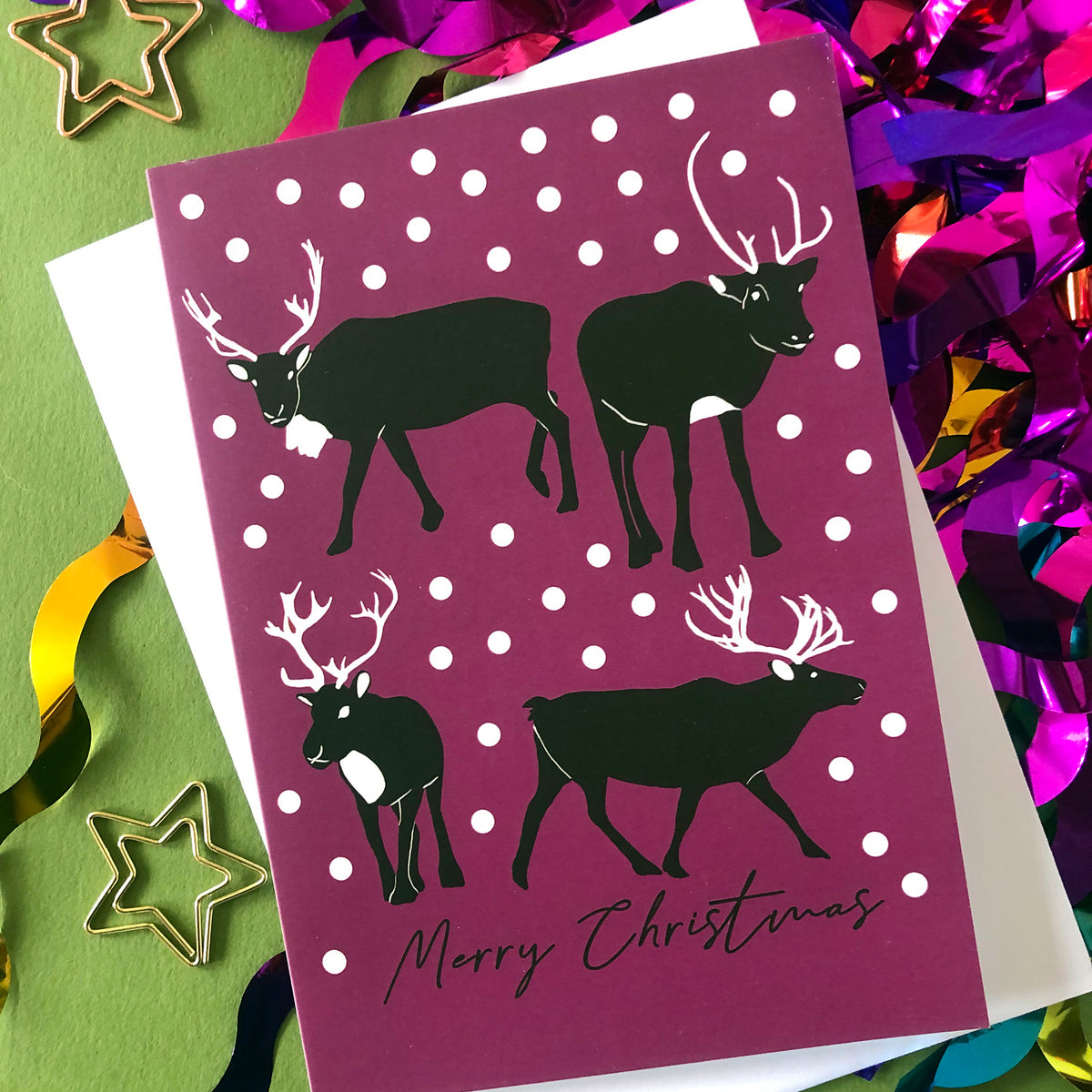 A Christmas greeting card featuring black reindeer silhouettes on a plum background with white snowballs and the words 'Merry Christmas' at the bottom. The card is styled on a green surface with white paper backing and colorful metallic tinsel.