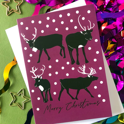 A Christmas greeting card featuring black reindeer silhouettes on a plum background with white snowballs and the words 'Merry Christmas' at the bottom. The card is styled on a green surface with white paper backing and colorful metallic tinsel.