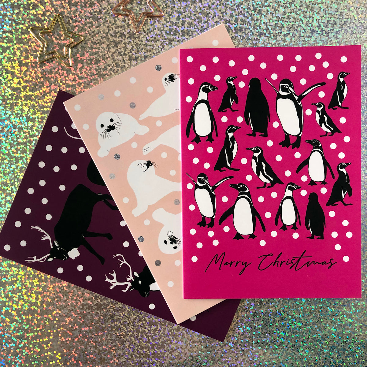 Three Christmas greeting cards fanned out on a glittery background. The front card is bright pink with penguin illustrations and the words 'Merry Christmas' in black script.