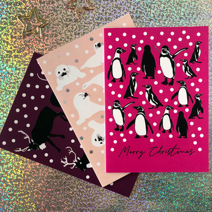Three Christmas greeting cards fanned out on a glittery background. The front card is bright pink with penguin illustrations and the words 'Merry Christmas' in black script.