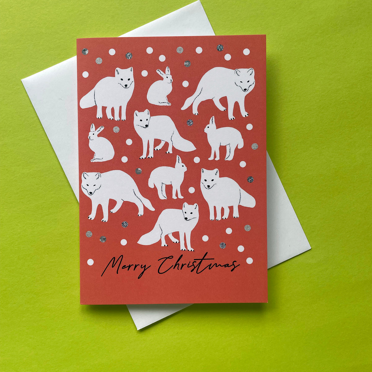 A Christmas greeting card featuring white illustrations of foxes and rabbits on a coral-red background with silver glitter accents, shown with a white envelope on a bright lime green surface.