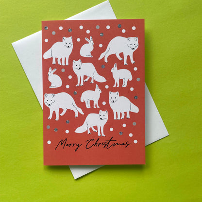A Christmas greeting card featuring white illustrations of foxes and rabbits on a coral-red background with silver glitter accents, shown with a white envelope on a bright lime green surface.