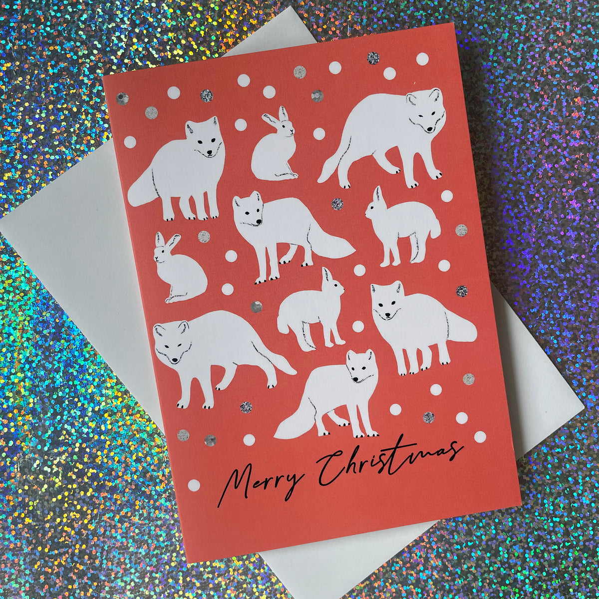 A Christmas greeting card featuring white illustrations of various foxes, rabbits, and a wolf on a coral-red background with silver glitter accents, resting on a white envelope over a colourful holographic glitter surface.