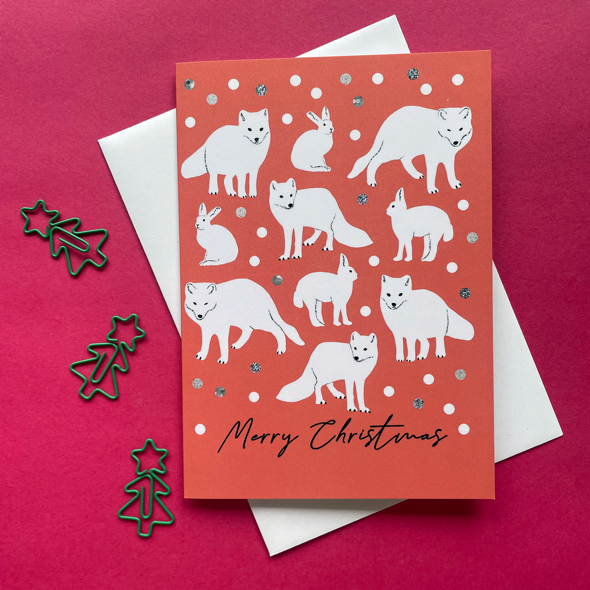 An orange Christmas greeting card featuring white illustrations of various foxes, wolves, and rabbits, with silver glitter accents and the text 'Merry Christmas' at the bottom. The card is displayed on top of a white envelope against a dark pink background, with three green metal Christmas tree cutouts on the left.