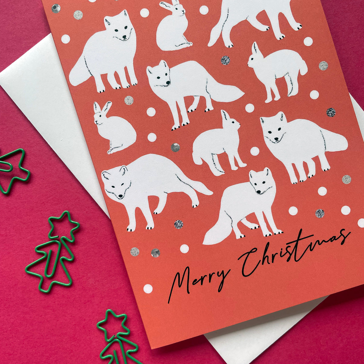 A Christmas greeting card with a terracotta orange-red background, decorated with white illustrations of foxes, a rabbit, and white snowballs, with 'Merry Christmas' written in black script. The card is layered over a white envelope on a dark pink background with green metal Christmas tree cookie cutters.