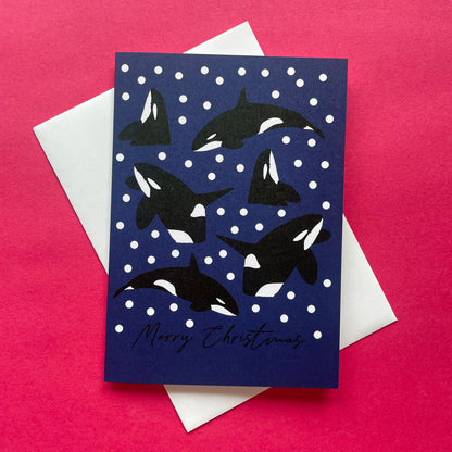 A Christmas greeting card featuring black and white killer whales on a dark blue background with white snowballs, layered over a white envelope on a bright pink background.