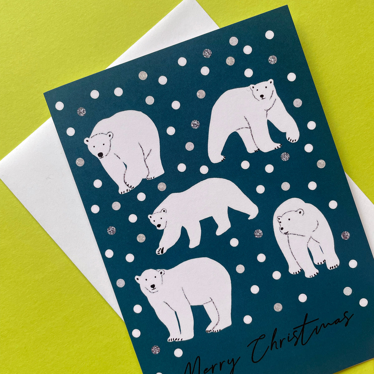 A Christmas greeting card featuring five white polar bear illustrations on a dark teal-blue background with white snowballs, shown with a white envelope on a bright yellow-green surface.