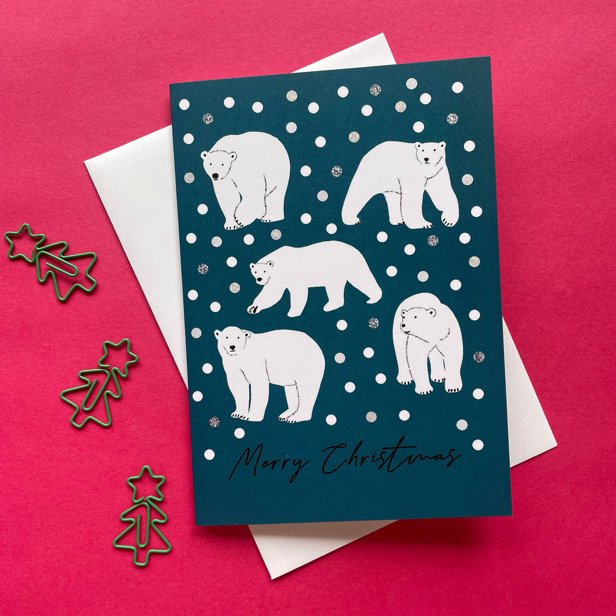 A Christmas greeting card featuring white polar bear illustrations on a dark teal background with white snowballs and the text 'Merry Christmas' at the bottom. The card is displayed on top of a white envelope against a bright pink background, with small metallic star and tree-shaped paperclips on the left.
