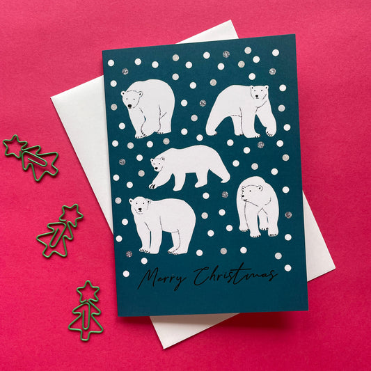 A Christmas greeting card featuring white polar bear illustrations on a dark teal background with white snowballs and the text 'Merry Christmas' at the bottom. The card is displayed on top of a white envelope against a bright pink background, with small metallic star and tree-shaped paperclips on the left.
