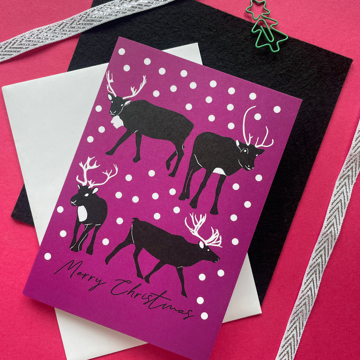 A Christmas greeting card with a purple background, white snowballs, and black reindeer silhouettes, layered over a black envelope and a white card on a pink background with ribbon accents.