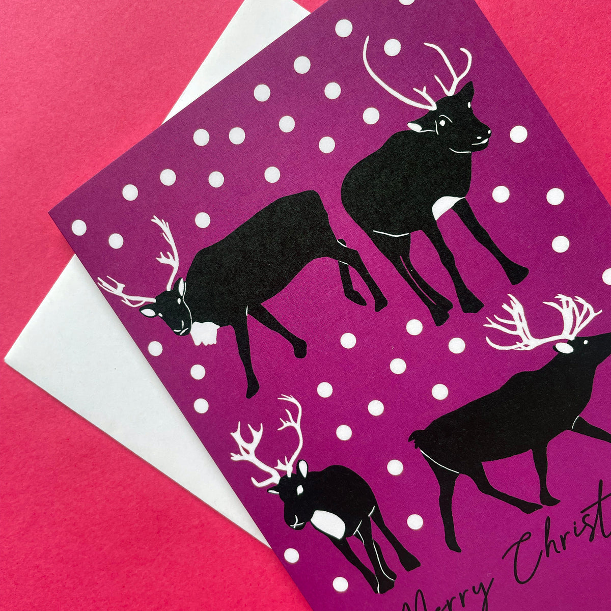 A Christmas greeting card featuring black reindeer silhouettes on a magenta-purple background with white snowballs, partially layered over a white envelope on a bright pink surface.