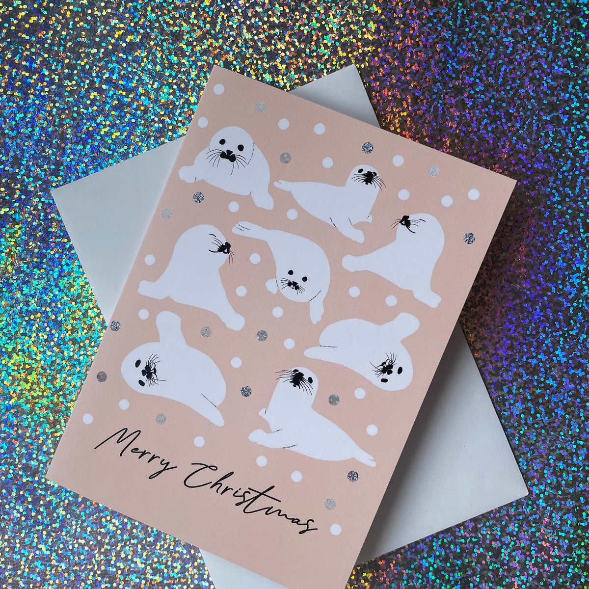 A Christmas greeting card featuring white seal illustrations on a pale peach background with silver glitter accents, shown with a white envelope on a colourful holographic glitter surface.