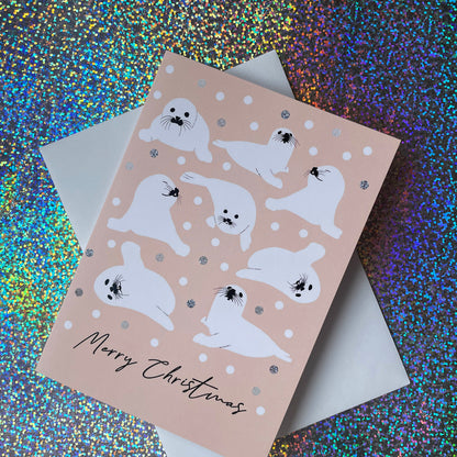 A Christmas greeting card featuring white seal illustrations on a pale peach background with silver glitter accents, shown with a white envelope on a colourful holographic glitter surface.