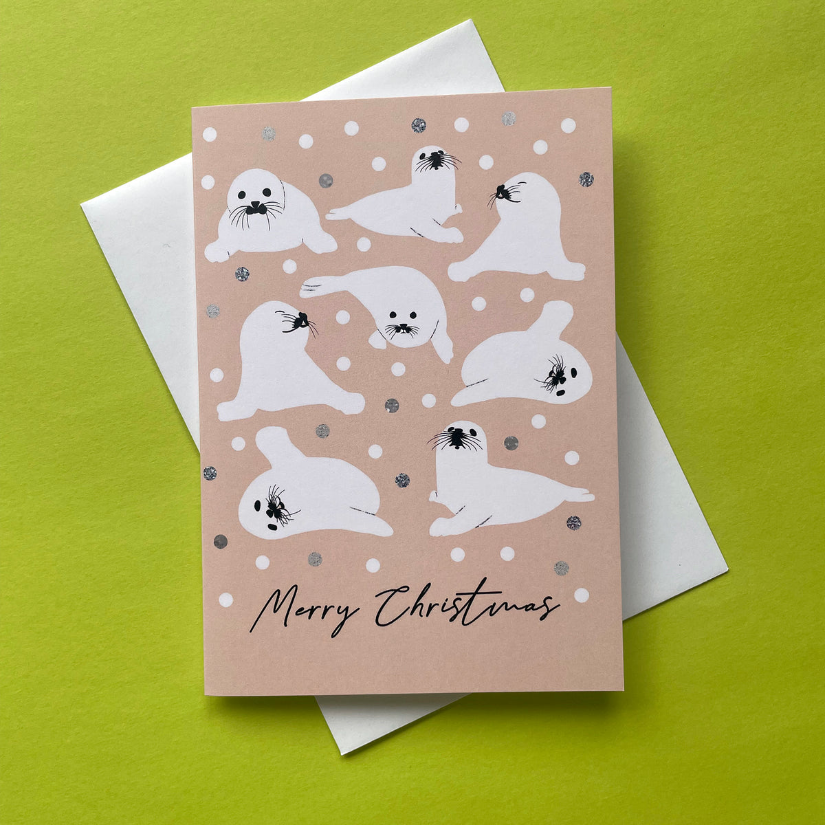 A Christmas greeting card featuring white seals on a peach background with snowballs,  layered over a white envelope on a bright lime green surface.