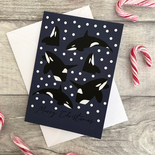 A Christmas greeting card featuring black and white killer whale illustrations on a dark navy blue background with white snowballs, layered over a white envelope. The card is styled on a grey wood-textured surface with candy canes in the corners.
