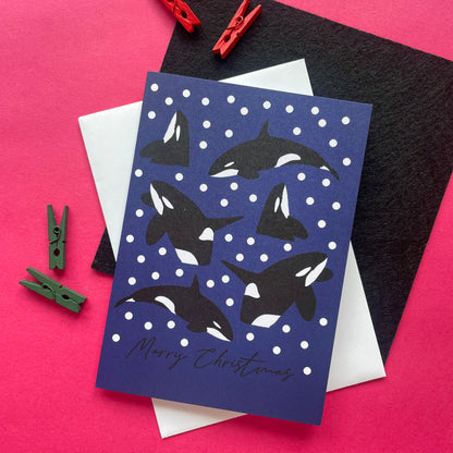A Christmas greeting card featuring black and white killer whale illustrations on a dark blue background with white snowballs, styled on a pink surface with red and green clothespins, a black textured sheet, and white envelopes.