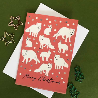 A Christmas greeting card with illustrations of white arctic foxes and rabbits on a coral-red background, placed on top of a white envelope. The card is photographed on a dark green surface with decorative metal star and tree cookie cutters around it.