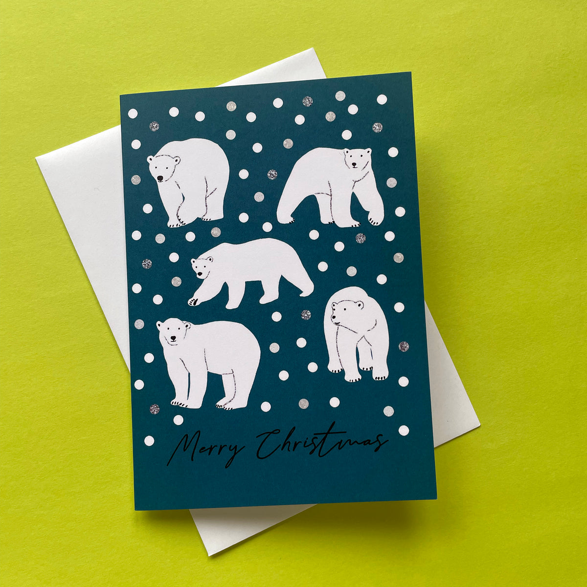 A Christmas greeting card featuring white polar bear illustrations on a dark teal background with sinowballs, layered over a white envelope on a bright yellow-green surface.