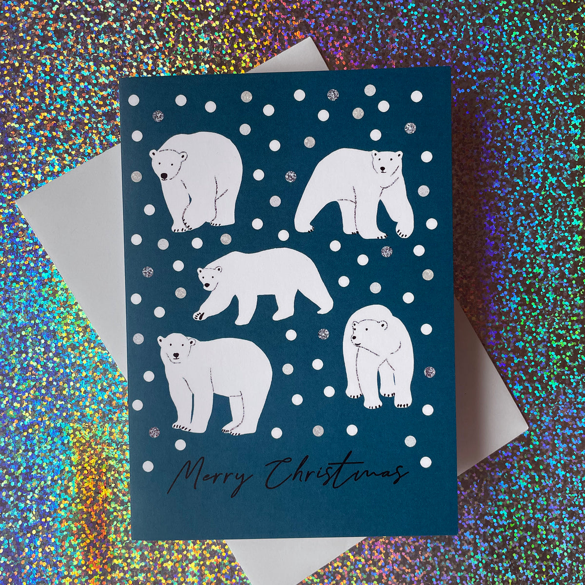 A dark teal Christmas greeting card featuring white polar bear illustrations on a snowy background with 'Merry Christmas' written at the bottom, layered over a white envelope against a holographic glitter backdrop.