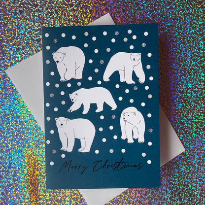 A dark teal Christmas greeting card featuring white polar bear illustrations on a snowy background with 'Merry Christmas' written at the bottom, layered over a white envelope against a holographic glitter backdrop.