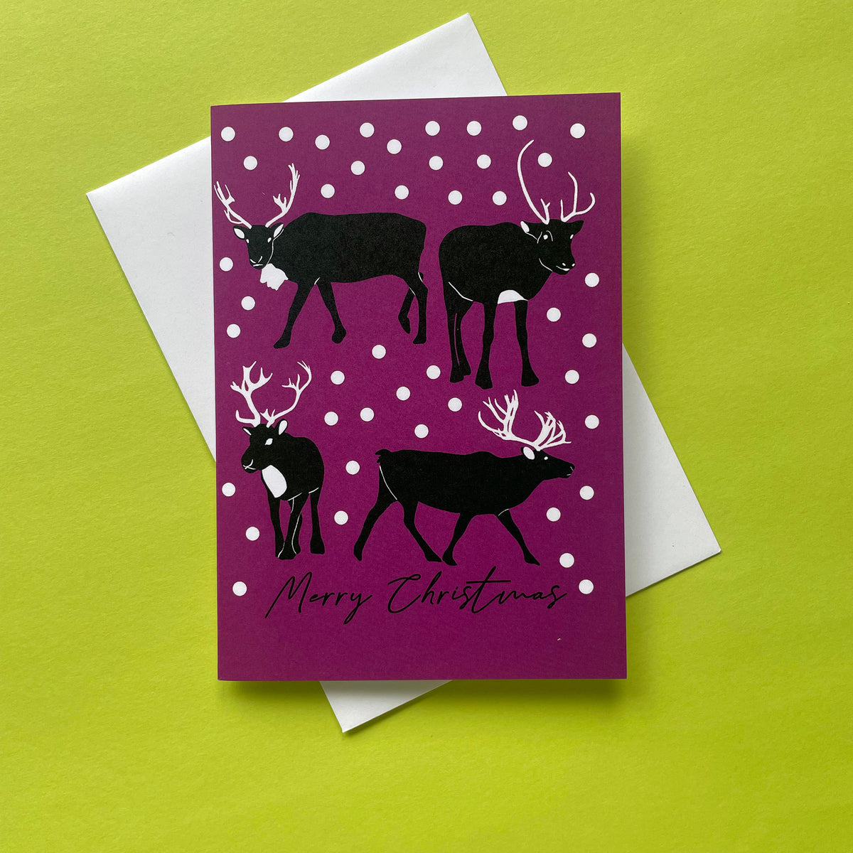 A Christmas greeting card featuring four black reindeer silhouettes on a deep purple background with white snowballs, placed on top of a white envelope against a bright yellow-green surface.