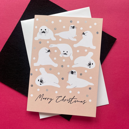 A Christmas greeting card featuring illustrated seals in winter poses on a warm beige background with silver and white snowballs layered over a white envelope and a black card on a bright pink surface.