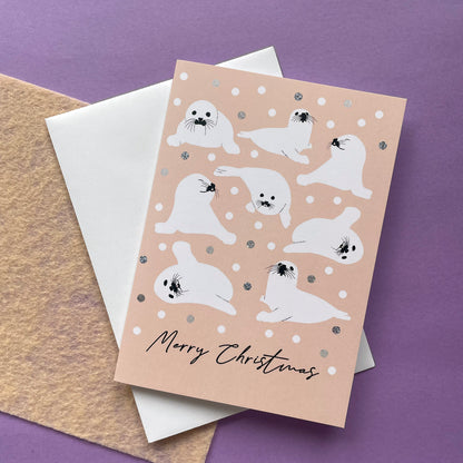 A Christmas greeting card featuring cute white seal illustrations on a peach background with snowballs, layered over a white envelope and a textured kraft paper sheet on a purple surface.