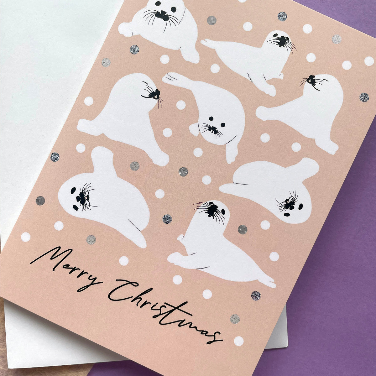 A Christmas greeting card featuring cute white seal illustrations on a peach background with silver glitter dots, layered over a white envelope. The card is angled on a purple surface.
