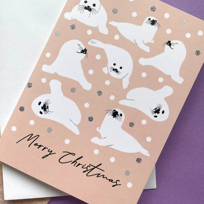 A Christmas greeting card featuring cute white seal illustrations on a peach background with silver glitter dots, layered over a white envelope. The card is angled on a purple surface.