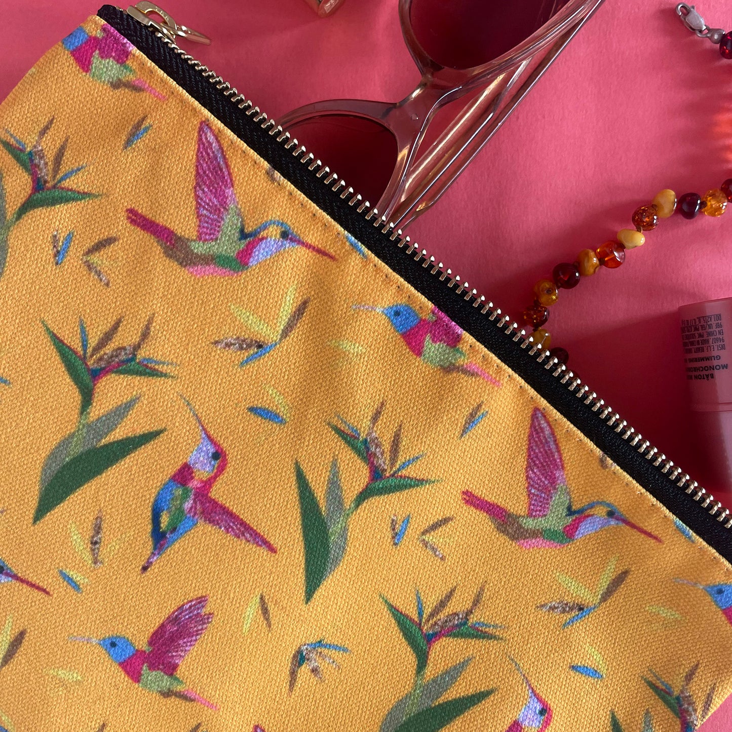 Image shows a bright yellow zip pouch with a colourful hummingbird and bird of paradise plant print.