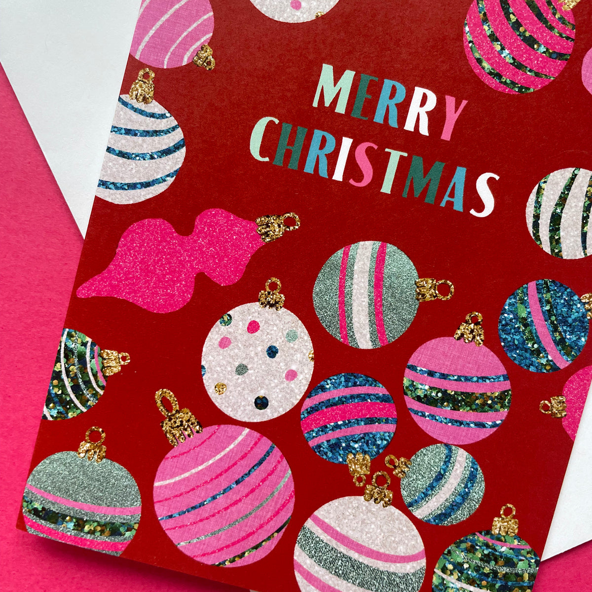 A red Christmas greeting card featuring a festive pattern of glittery and striped ornaments, with the words 'MERRY CHRISTMAS' printed in colourful lettering. The card is photographed at an angle on a bright pink surface with a white envelope partially visible.