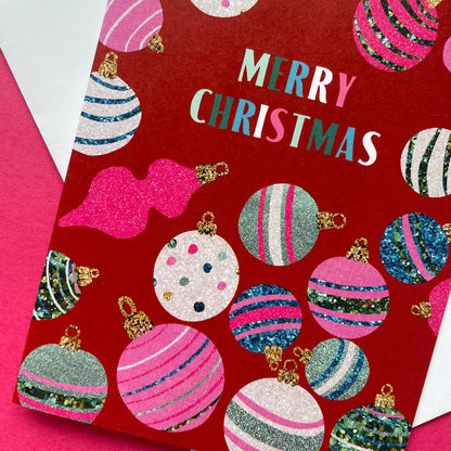A red Christmas greeting card featuring a festive pattern of glittery and striped ornaments, with the words 'MERRY CHRISTMAS' printed in colourful lettering. The card is photographed at an angle on a bright pink surface with a white envelope partially visible.